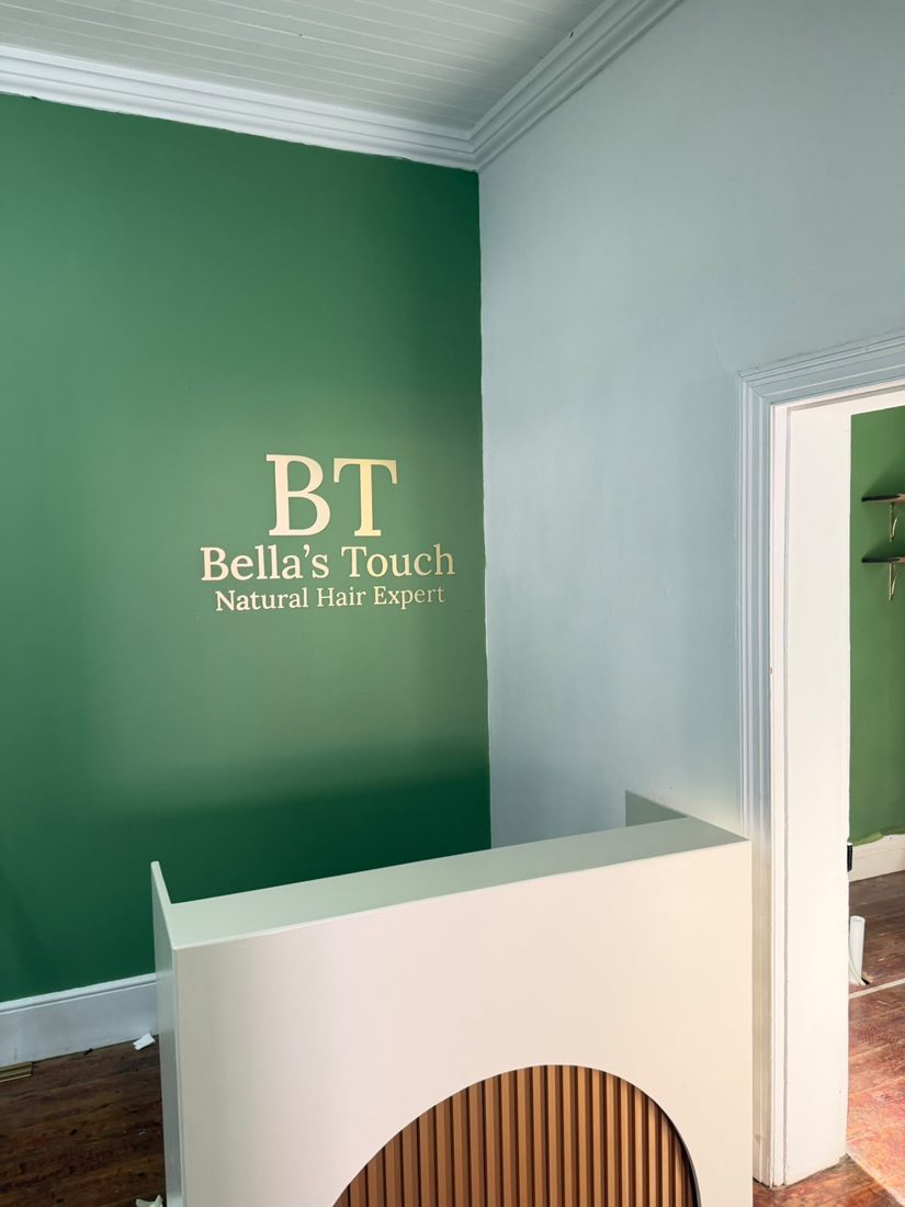 Gold raised-letter Bella's Touch logo sign on a green feature wall above a white reception desk